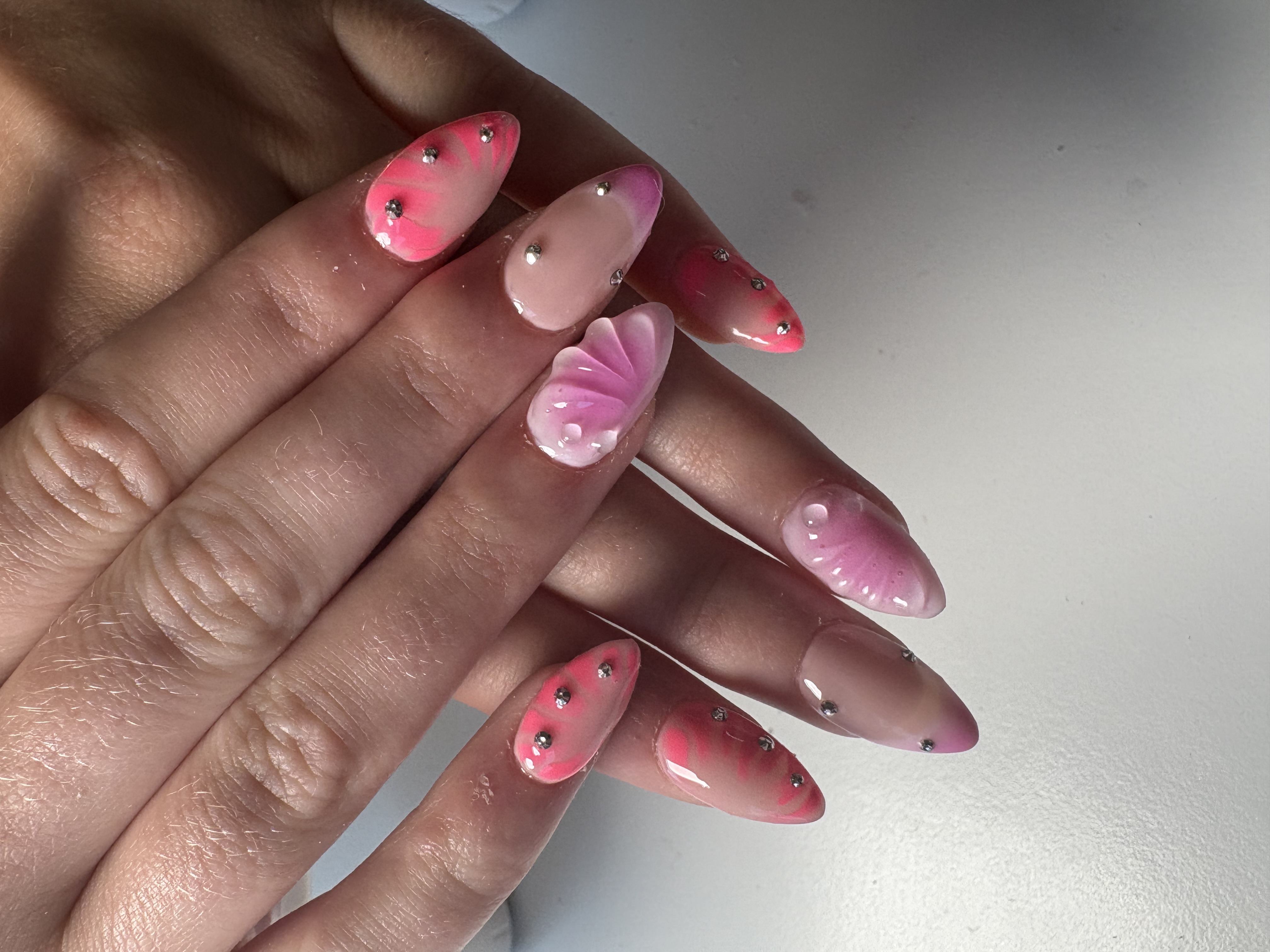 Pink almond nails with tie-dye swirls, a 3D sculpted floral accent, and rhinestones