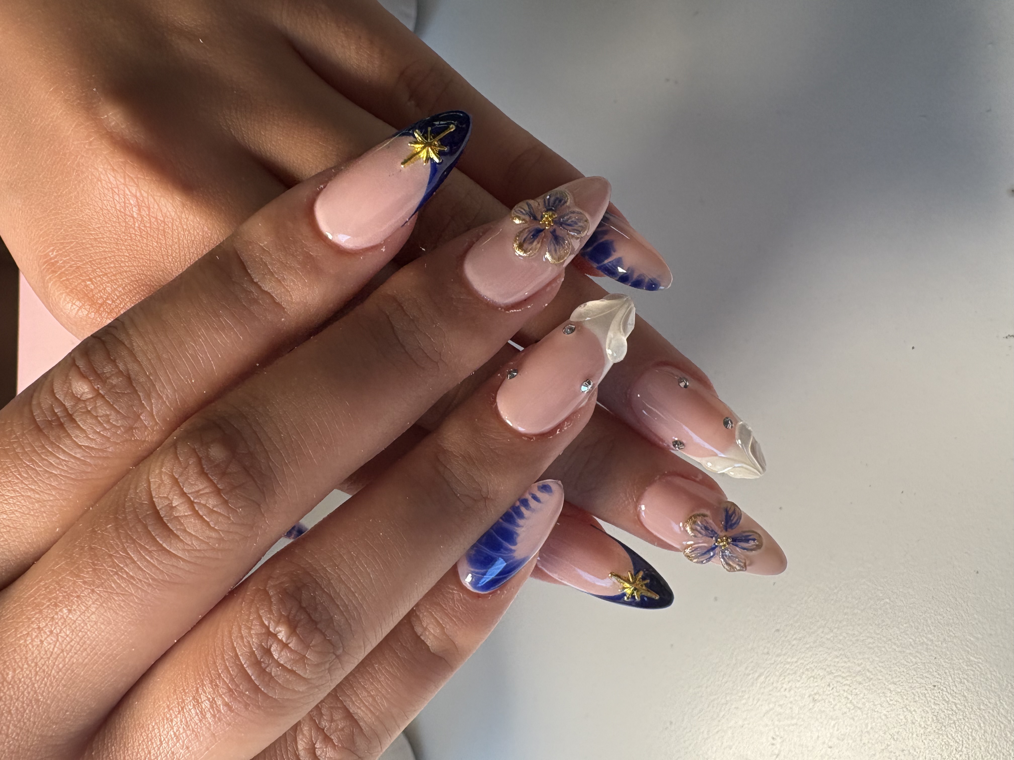 Nude almond nails with blue tie-dye accents, 3D gold flowers, and chrome star tips