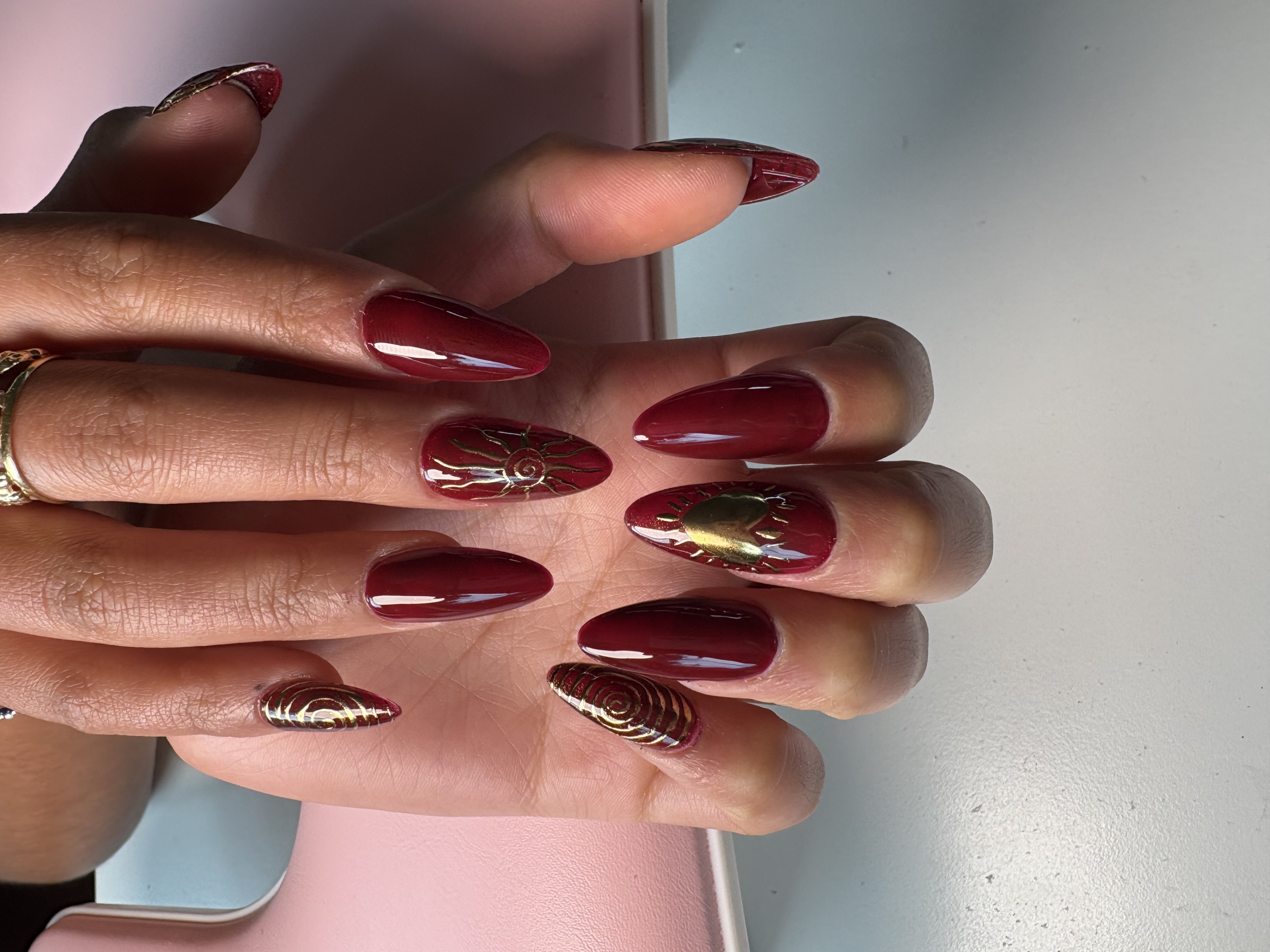 Deep red almond nails with engraved gold sun-spiral and heart linework accents