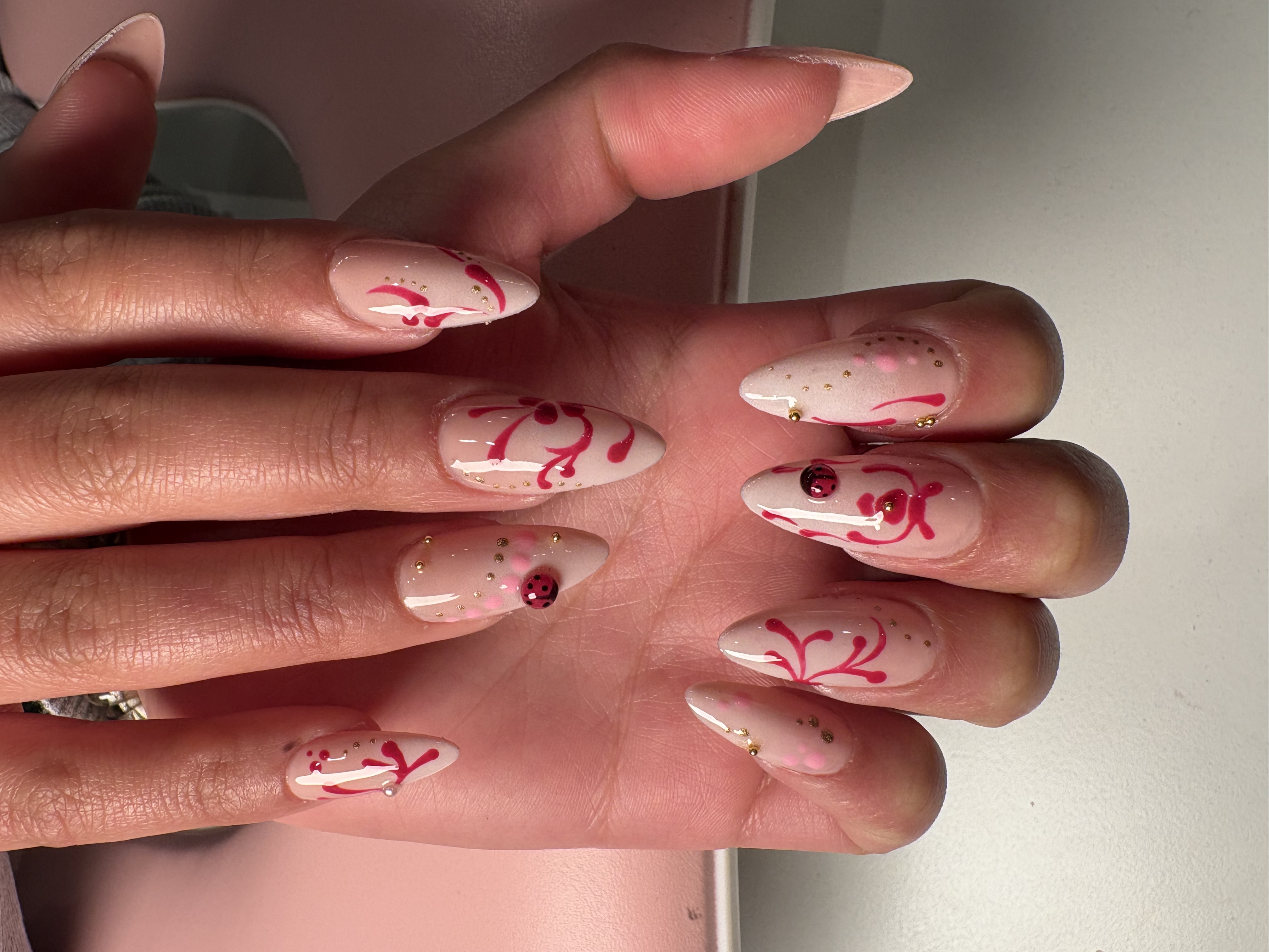 Nude stiletto nails with hand-painted pink vine art, gold studs, and ladybug charms