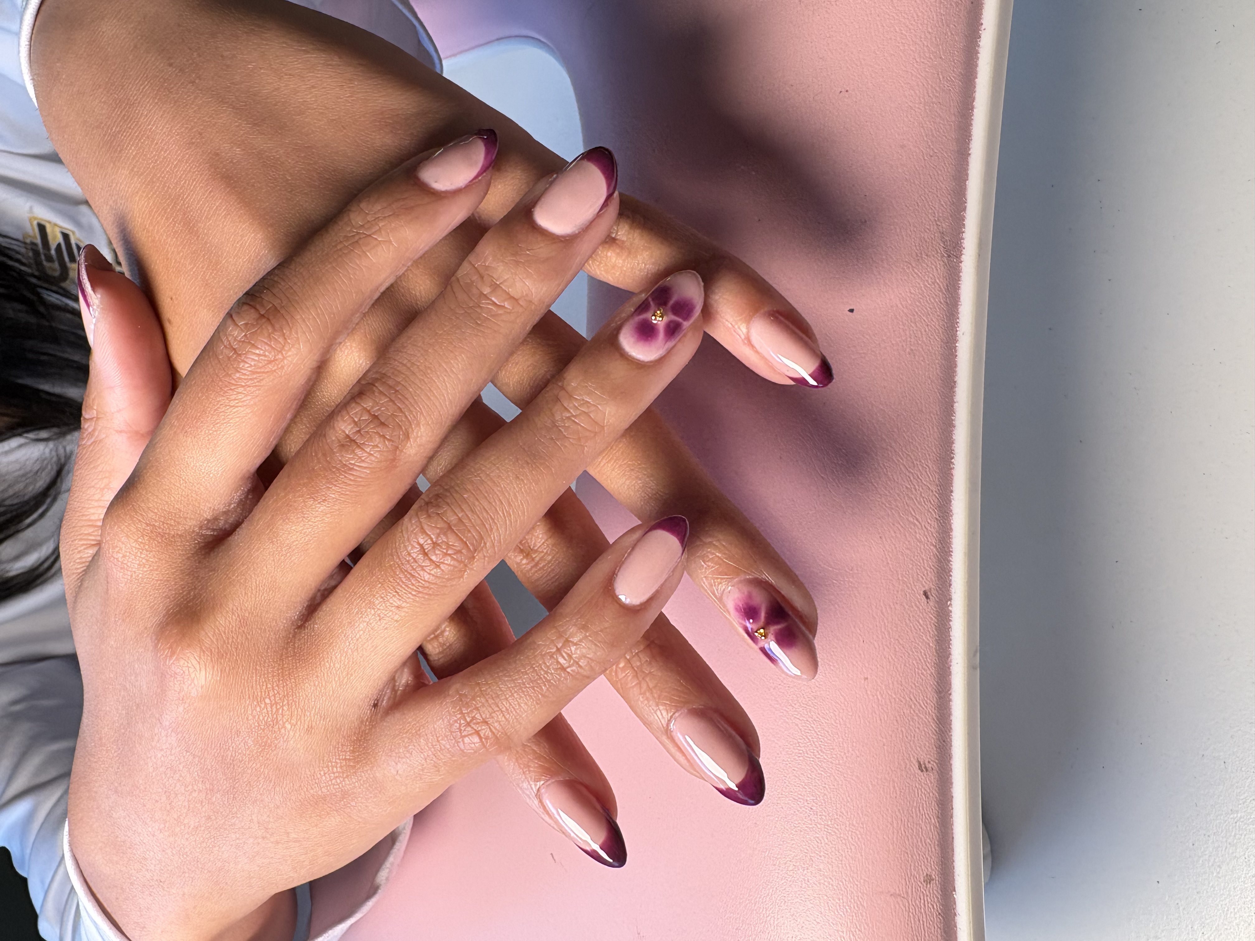 Nude oval nails with a burgundy French tip and a purple flower accent