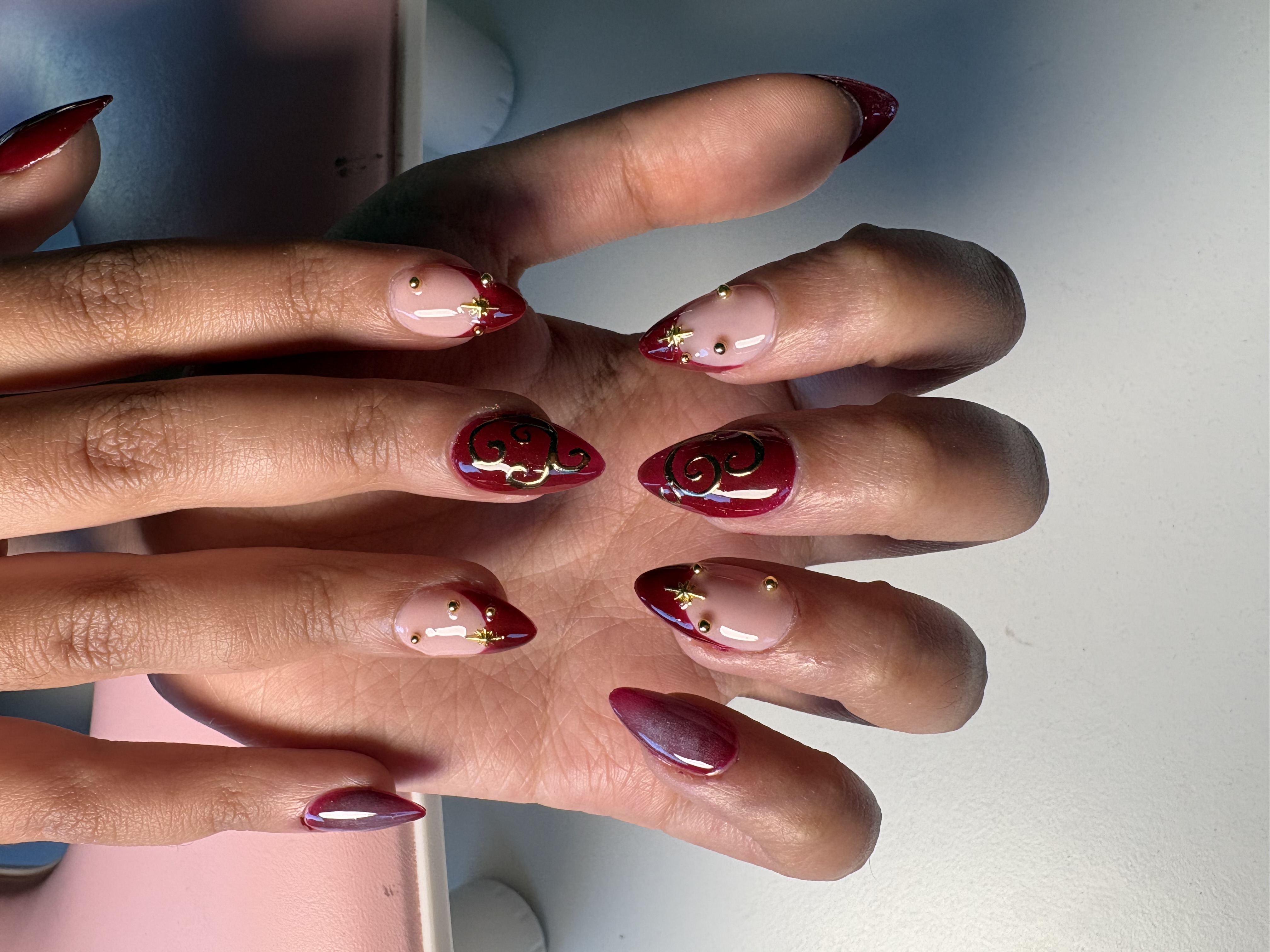 Burgundy almond nails with gold starburst charms and intricate gold swirl linework accents