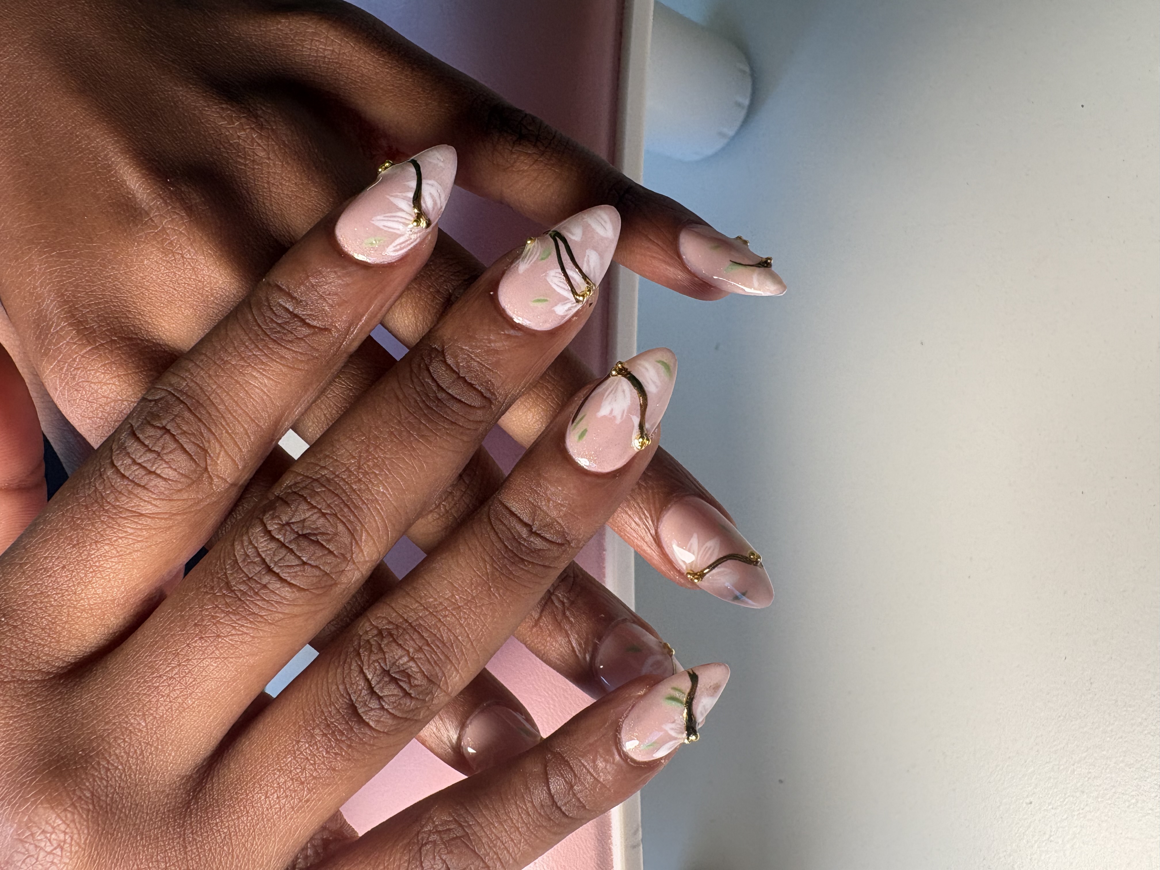 Nude almond nails with hand-painted white flowers, green leaves, and thin gold vines