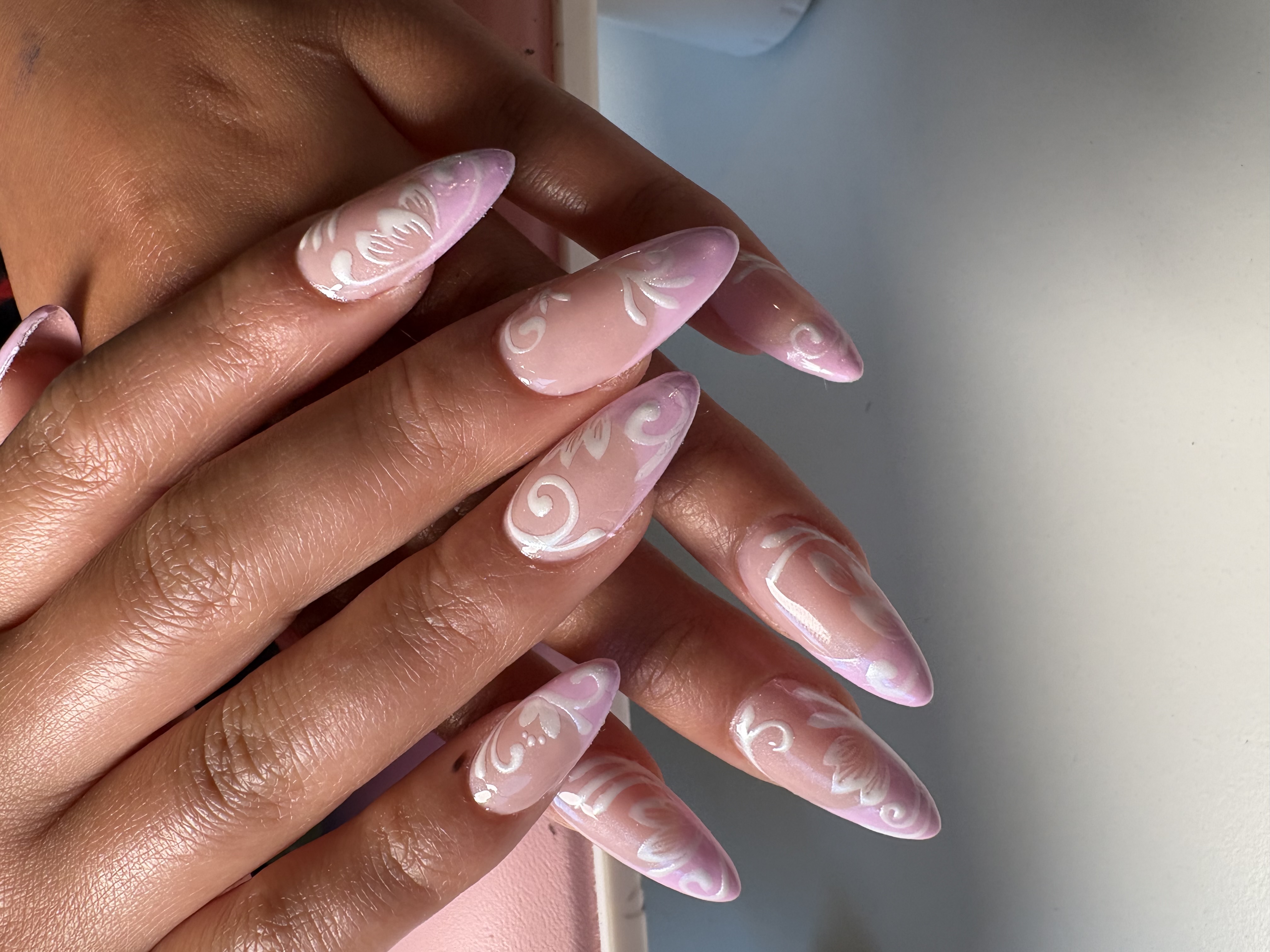 Nude almond nails with intricate white lace-like floral scrollwork covering most nails